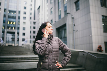 Fototapeta premium young girl talking on mobile phone in courtyard business center. girl with long dark hair dressed in winter jacket in cold weather speaks on phone on background buildings made of glass and concrete