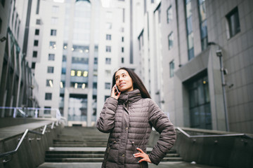Fototapeta premium young girl talking on mobile phone in courtyard business center. girl with long dark hair dressed in winter jacket in cold weather speaks on phone on background buildings made of glass and concrete