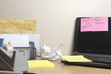 Out of office, back soon message left on a laptop on a messy office desk 