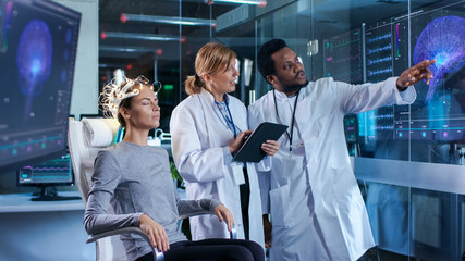 Woman Wearing Brainwave Scanning Headset Sits in a Chair while Two Scientists Supervise and Look at Data. In the Modern Brain Study Laboratory Monitors Show EEG Reading and Brain Model.