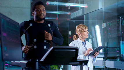 Male Athlete Runs on a Treadmill with Electrodes Attached to His Body while Sport Scientist Holds Tablet and Supervises EKG Status. In the Background Laboratory with High-Tech Equipment. Dark Ambient.