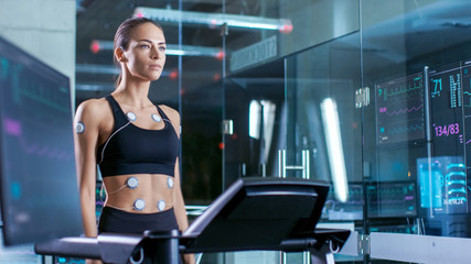 Fototapeta na wymiar Beautiful Woman Athlete Wearing Sports Bra with Electrodes Connected to Her, Walks on a Treadmill in a Sports Science Laboratory. In the Background Laboratory. Medium Shot.