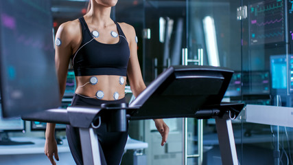 Beautiful Woman Athlete with Electrodes Attached to Her, Running on a Treadmill in a Sports Science Laboratory. In the Background Lab with Monitors Showing EKG data.