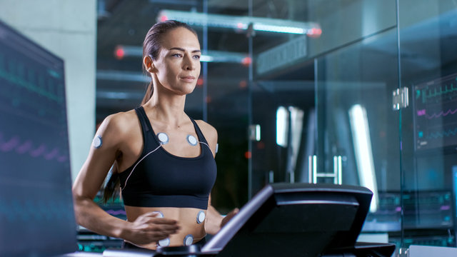 Beautiful Woman Athlete with Electrodes Attached to Her, Running on a Treadmill in a Sports Science Laboratory. In the Background Lab with Monitors Showing EKG data.