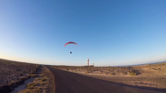 Motor paraglider in deep flight mode at sunset passing a lighthouse.