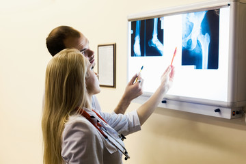 A group of mixed-race physicians discusses the diagnosis behind a laptop in the office. In the background, the asian professor explains to students the physicians anatomy