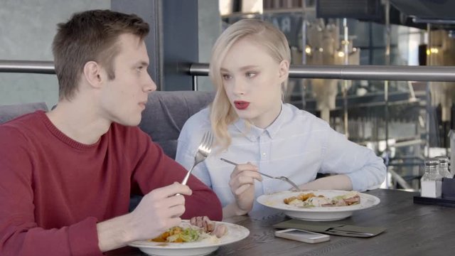 Pair of colleagues are having lunch together in stylish restaurant, slow motion. Young man in bordo jumper and blond woman in blue shirt are sitting on velvet sofa at the wooden table, eating salad