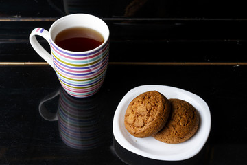 on a black background two oatmeal cookies lie on a white plate, next is a large striped mug with tea