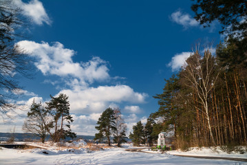 Przydrożna kapliczka na skraju lasu w zimowej szacie oświetlone porannym słońcem z niebieskim niebem z chmurami - obrazy, fototapety, plakaty
