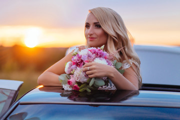 Dreamy bride stands with wedding bouquet over a car with open hood