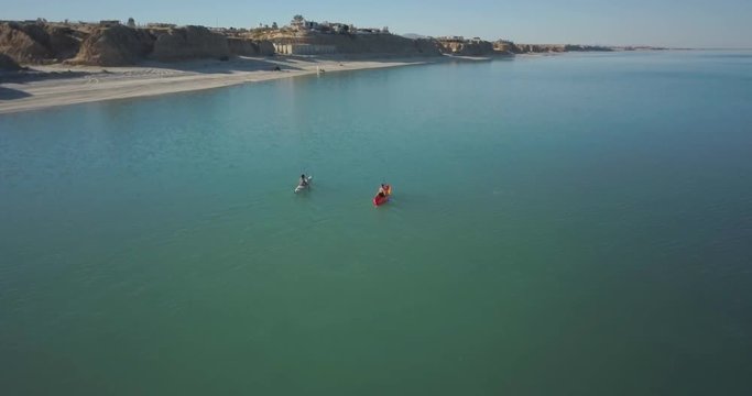 Aerial camera follows behind two kayakers paddling along a sandy beach and cliffs.