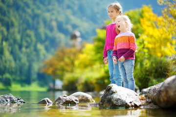 Adorable sisters playing by Konigssee lake in Germany on warm summer day. Cute children having fun feeding ducks and throwing stones into the lake.