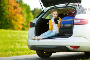 Adorable little girl ready to go on vacations with her parents. Kid sitting in a car trunk and reading her ebook.