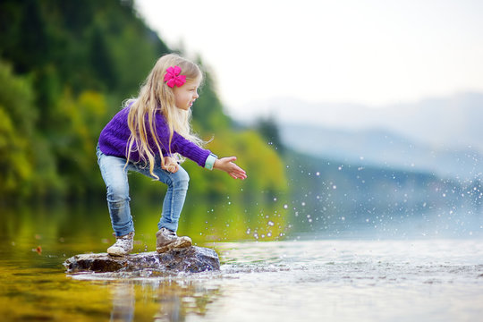 Adorable Girl Playing By Hallstatter See Lake In Austria On Warm Summer Day. Cute Child Having Fun Splashing Water And Throwing Stones Into The Lake.