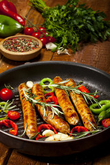 Fried sausages with herbs, spices and vegetables in a pan. On wooden background.