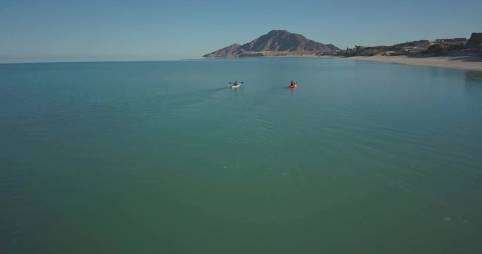 Aerial view of two kayakers paddling in a sea toward a mountain in the distance.