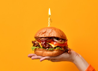 Woman hands hold big burger barbeque sandwich with beef and lit candle for birthday party on yellow - obrazy, fototapety, plakaty