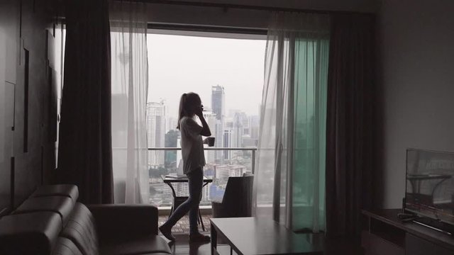 Young female freelancer or businessman ceo talking on the phone, negotiating the panoramic windows and terrace in luxury apartment on business trip. Silhouette of woman using smartphone.