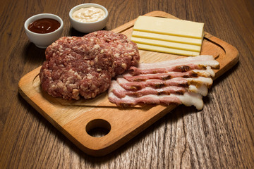 Ingredients for cooking burgers. Raw ground beef meat cutlets on wooden chopping board, cheese, bacon,  over wooden background