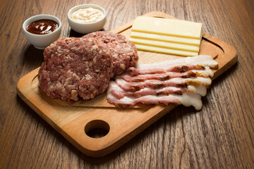 Ingredients for cooking burgers. Raw ground beef meat cutlets on wooden chopping board, cheese, bacon,  over wooden background
