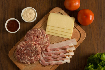 Ingredients for cooking burgers. Raw ground beef meat cutlets on wooden chopping board, cheese, bacon,  over wooden background