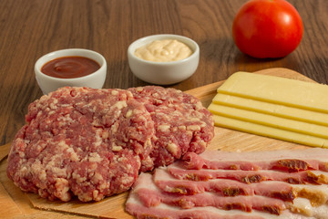 Ingredients for cooking burgers. Raw ground beef meat cutlets on wooden chopping board, cheese, bacon,  over wooden background