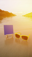 Sunglasses on sand with blue board at sunset, beach and ocean in the background
