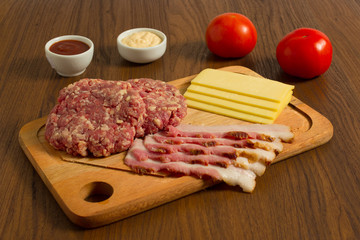 Ingredients for cooking burgers. Raw ground beef meat cutlets on wooden chopping board, cheese, bacon,  over wooden background