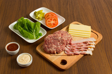 Ingredients for cooking burgers. Raw ground beef meat cutlets on wooden chopping board, tomatoes, greens, pickles, ketchup, cheese,  over wooden background 
