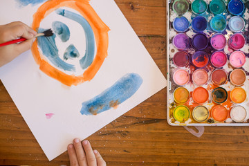 Child painting orange and blue smiley face with watercolors on table
