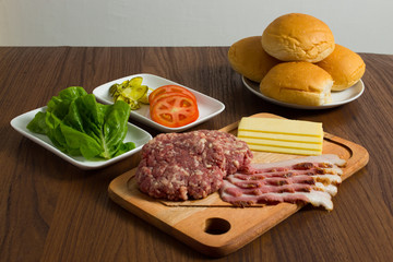Ingredients for cooking burgers. Raw ground beef meat cutlets on wooden chopping board, tomatoes, greens, pickles, ketchup, cheese,  over wooden background 