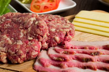Ingredients for cooking burgers. Raw ground beef meat cutlets on wooden chopping board, tomatoes, greens, pickles, ketchup, cheese,  over wooden background 