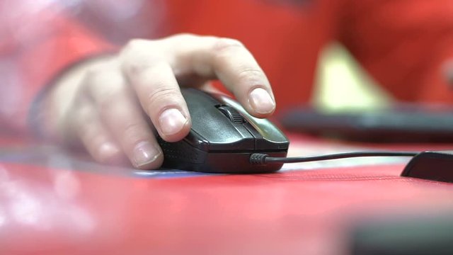 Hand of male gamer, who is holding black mouse, close up. Man in red shirt is playing on computer and using the device on the table to cooperate with it by pushing buttons by his fingers.