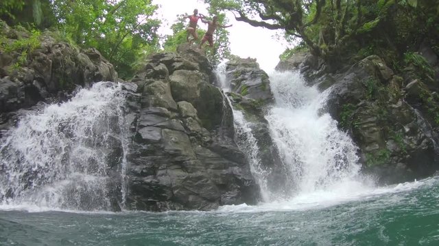SLOW MOTION Young couple on a summer adventure jump into water from a ledge while holding hands. Man and woman on an exciting date in the outdoors. Newlyweds enjoying their tropical honeymoon in Fiji