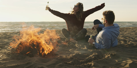 Loving Young Couple Sitting On The Beach beside Campfire drinking beer