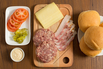 Ingredients for cooking burgers. Raw ground beef meat cutlets on wooden chopping board, tomatoes, greens, pickles, ketchup, cheese,  over wooden background - Top View