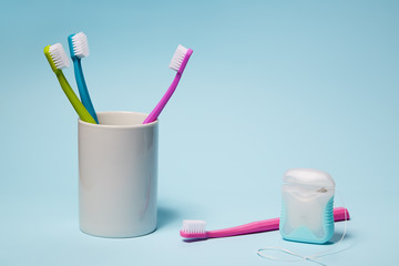Colorful toothbrushes in mug and dental floss on light blue background. 