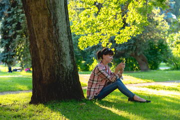 young woman sits under a tree with a phone in hands in the summe