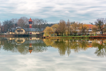 Fototapeta na wymiar Beautiful lake with mirror reflections in clear water on cloudy day. Tranquil landscape with lake, houses, cloudy sky, and trees reflected symmetrically in the water. Nyiregyhaza, Hungary