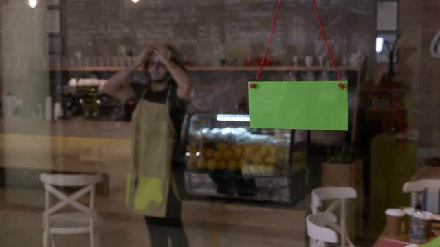 Small business owner of coffee shop starting the day at work by putting his apron on and flipping green screen sign on the door