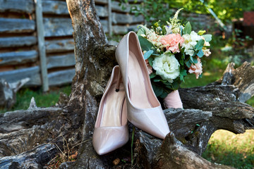 Close up of beautiful beige shiny bride shoes and wedding bouquet of pink and white roses on wooden stump outdoors. Wedding morning preparation. Wedding bride accessories. Bridal bouquet and shoes