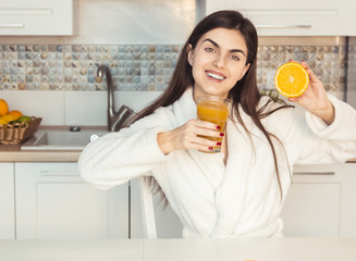 Fototapeta premium Beautiful girl wears white bathrobe with glass of orange juice and fruit, kitchen morning sunset view