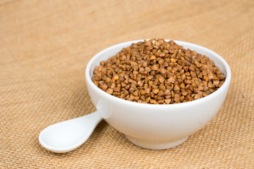 dry buckwheat heap in white bowl with spoon over sackcloth