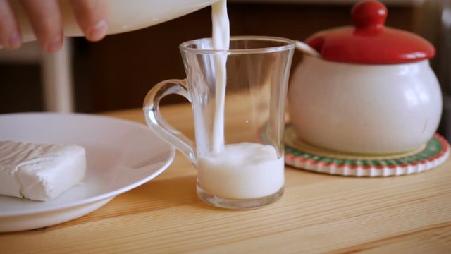 A man pours milk into a glass mug on a wooden table. Cottage cheese on a plate. Slow Motion.