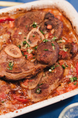 Casserole dish with braised veal shanks slow cooked or baked in the oven until tender in tomatoes, onions, carrots, celery and meat juice, so called Italian Ossobuco Milanese, served with gremolata