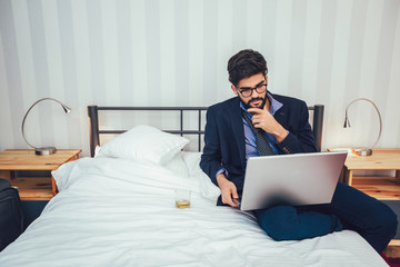 Working in hotel room. Confident young businessman in suit and tie working on laptop while sitting on the bed in hotel room