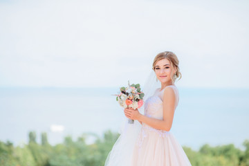 Charming bride in peach dress smiles posing before a great sea view in a sunny day