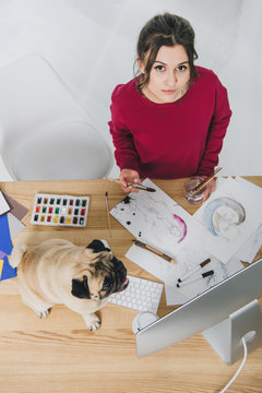 Attractive young girl working on illustrations with cute pug on working table with computer