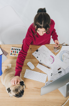 Young woman cuddling pug dog while working on designs by table