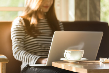 Cup of coffee in coffee shop with woman work with laptop in background in the morning